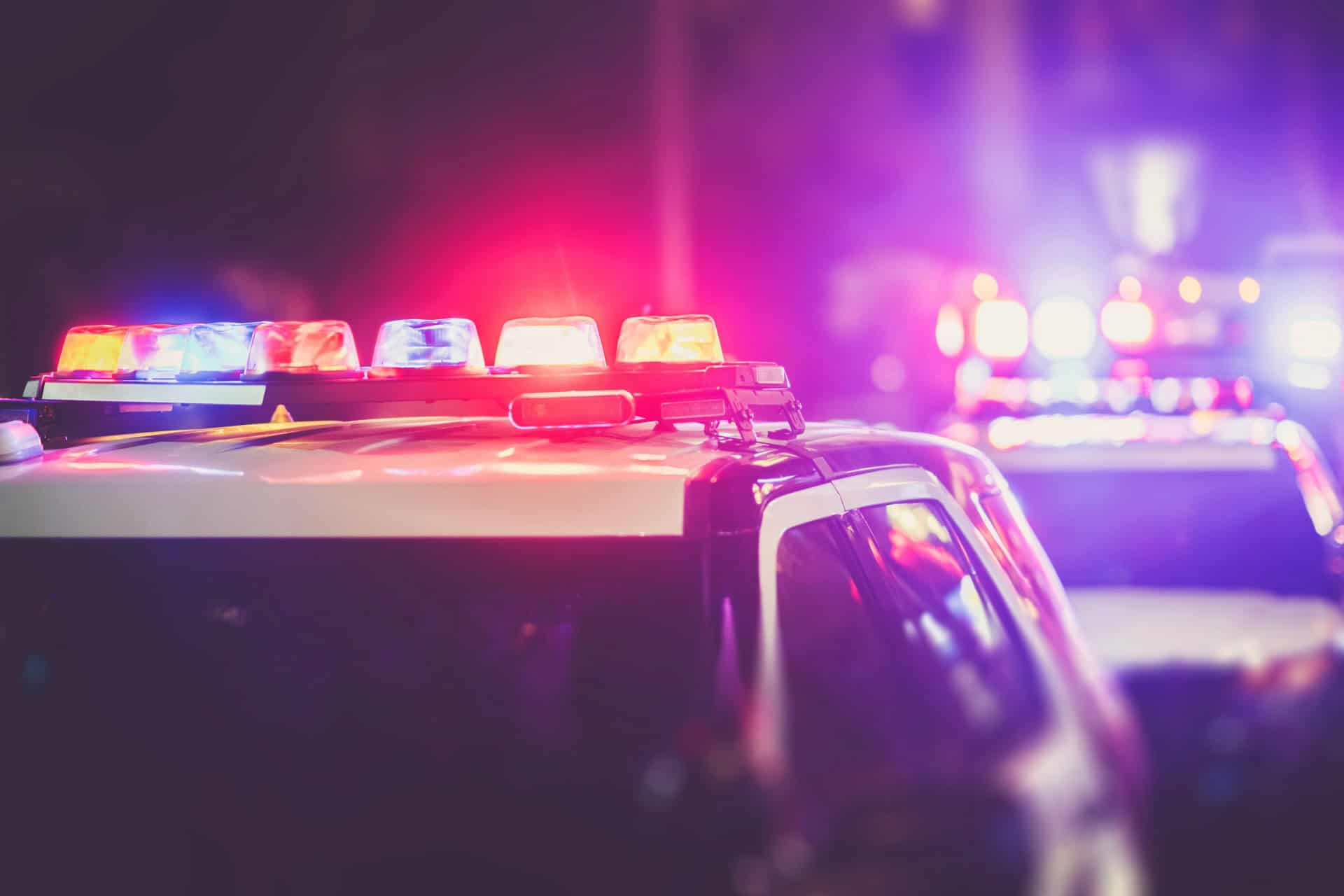 Police cars with flashing red and blue lights are parked on a street at night, with a blurred background of additional emergency vehicles.