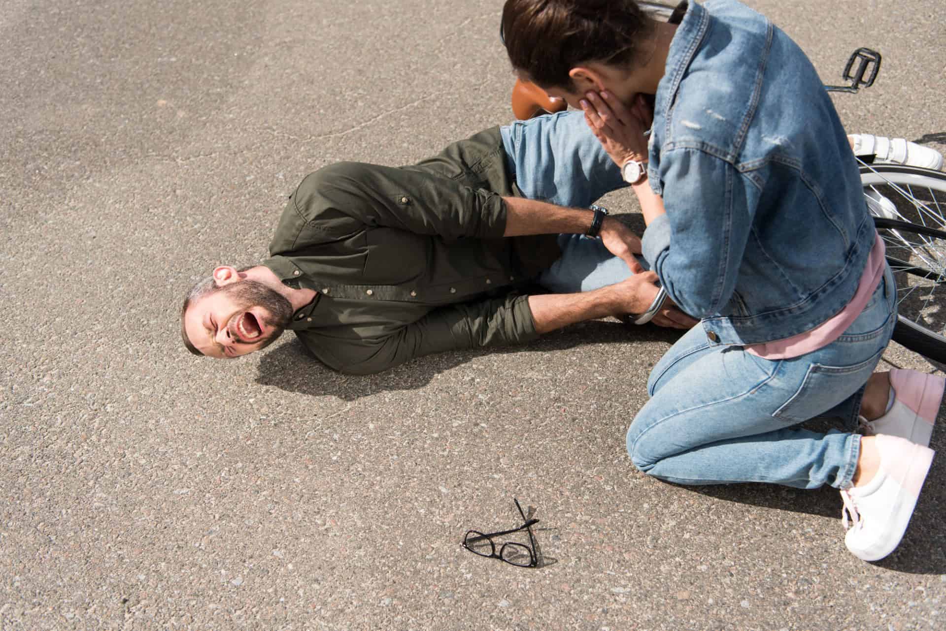 A man lies on the ground in pain next to a bicycle, holding his leg, while a woman kneels beside him, appearing concerned. Glasses are also on the ground nearby.