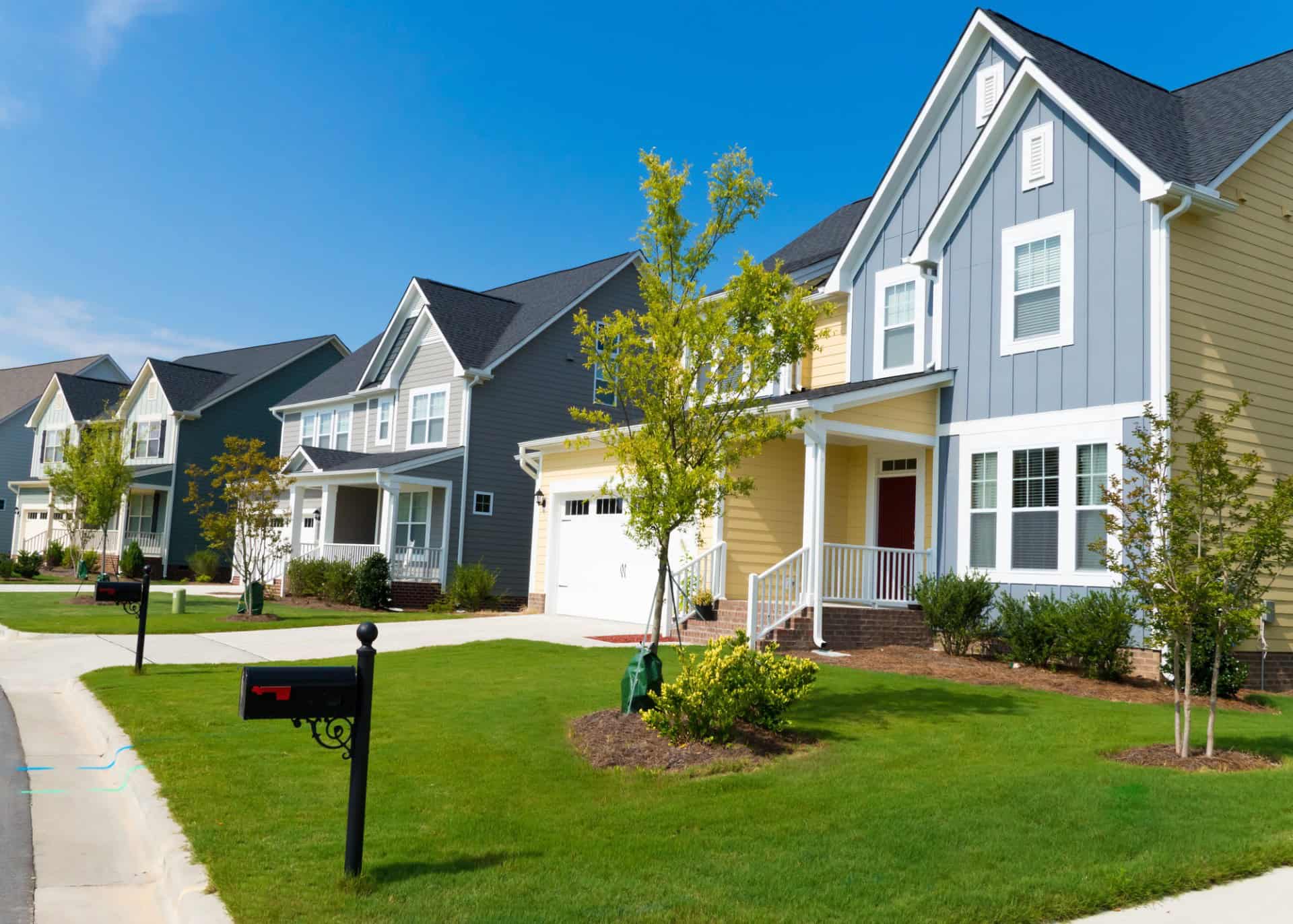 Row of modern suburban houses with well-kept lawns, mailboxes at the curb, and a clear blue sky in the background.